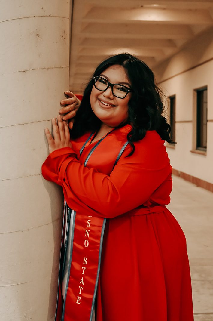 young woman standing next to a column, wearing a red dress and graduation sash