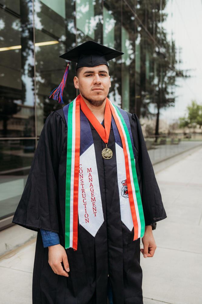 Pablo Cabrera is posed outside of the Fresno State library. He is wearing a cap and gown and graduation sashes and medals.
